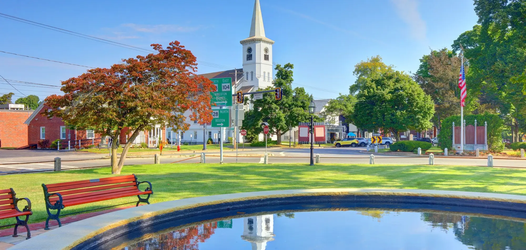 Central Square in Bridgewater on a clear morning. The church clock tower stands behind the intersection, with route signs for 104 east and 28 north to Boston, a flag and war memorial to the right, and benches beside the fountain in the foreground.